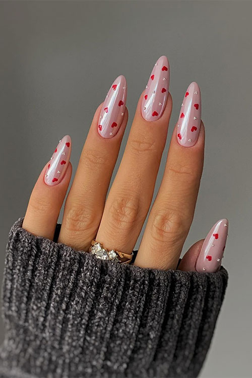 Glossy nude almond-shaped gel nails adorned with tiny white pearls and red heart shapes Glossy nude almond-shaped gel nails adorned with tiny white pearls and red heart shapes