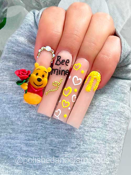 Long nude acrylic valentines nails 2023 with white and red hearts, rhinestones, yellow glitter, and bee mine words