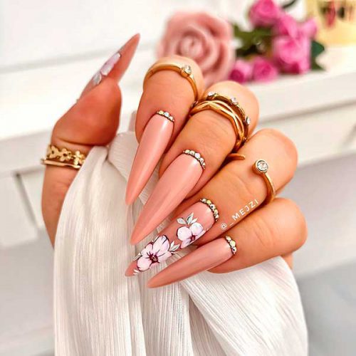 Long stiletto nude nails with pink floral nail art on two accent nails adorned with rhinestones above cuticles