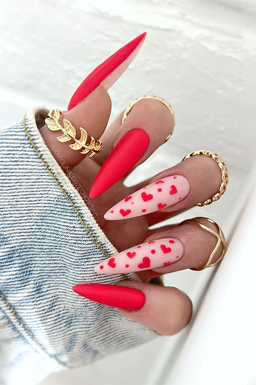 Stiletto matte red Valentine’s Day nails with two nude pink accent nails adorned with tiny red heart shapes Stiletto matte red Valentine’s Day nails with two nude pink accent nails adorned with tiny red heart shapes
