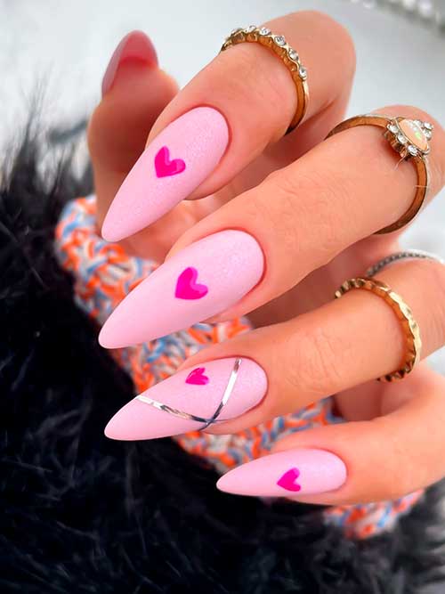 Long almond-shaped glitter pastel pink valentine’s day nails with hot pink hearts and silver crossed strips on an accent nail