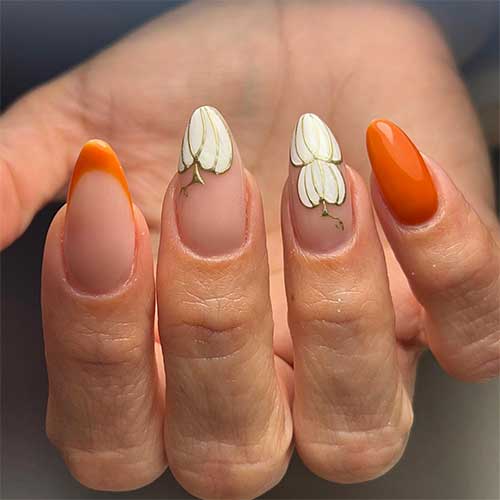 Almond shaped burnt orange nails with white pumpkins on two accent nude nail tips.