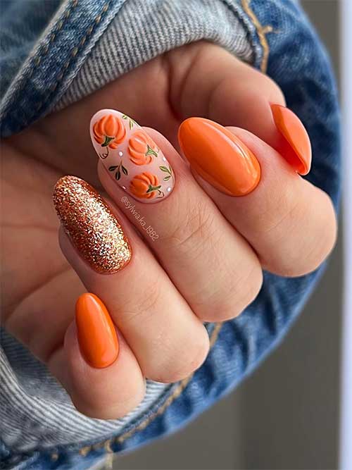 Cute orange almond-shaped nails with a gold glitter accent nail and orange pumpkins with green leaves on a nude accent nail