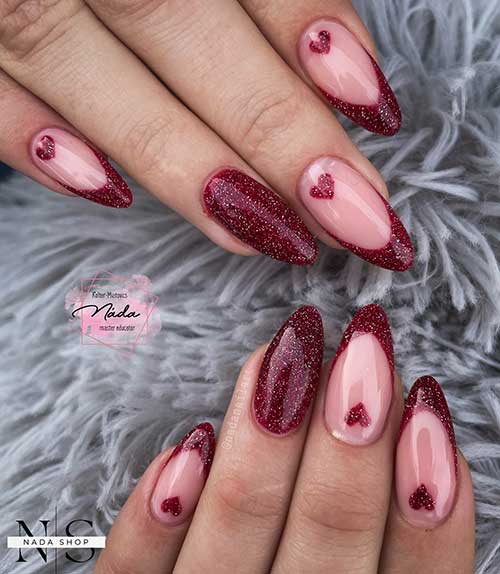 Dark red glitter French nails over nude base color and adorned with a heart shape on each nail above the cuticle area