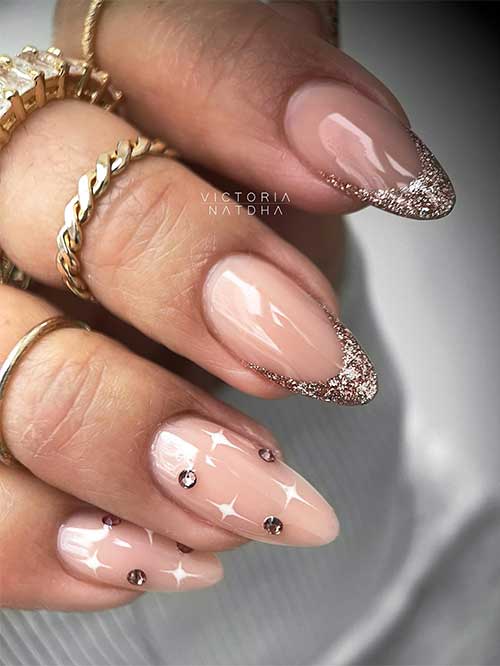 Long almond shaped rose gold glitter French New Year’s nails with two accent nude nails adorned with rhinestones and white stars.