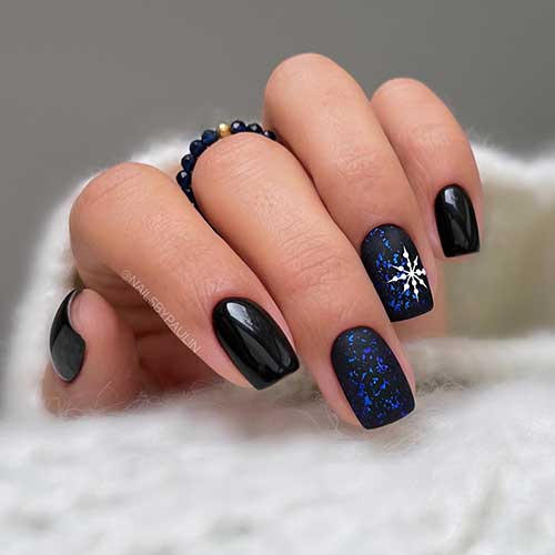 Short black winter nails with two accent black nails with blue flakes and a big white snowflake on the ring fingernail