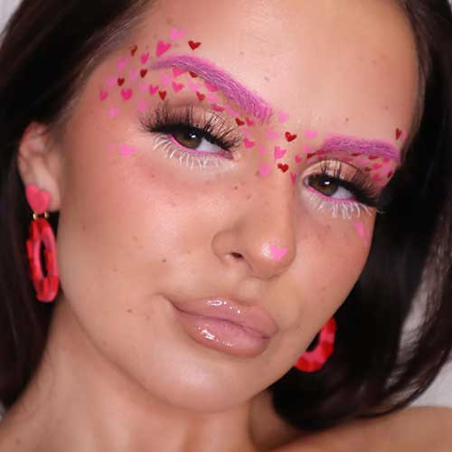 Scattered red and pink heart Valentine's Day eye makeup with pink bleached eyebrows, nude eyeshadow, and glossy nude lips