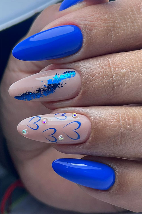 Glossy royal blue nails with two nude accent nails adorned with blue foil, blue outlined heart shapes, and some pearls