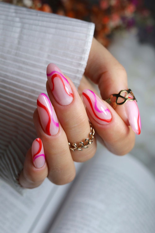Long almond-shaped glossy nude nails using rubber base milky rose and adorned with red and hot pink swirls on each nail