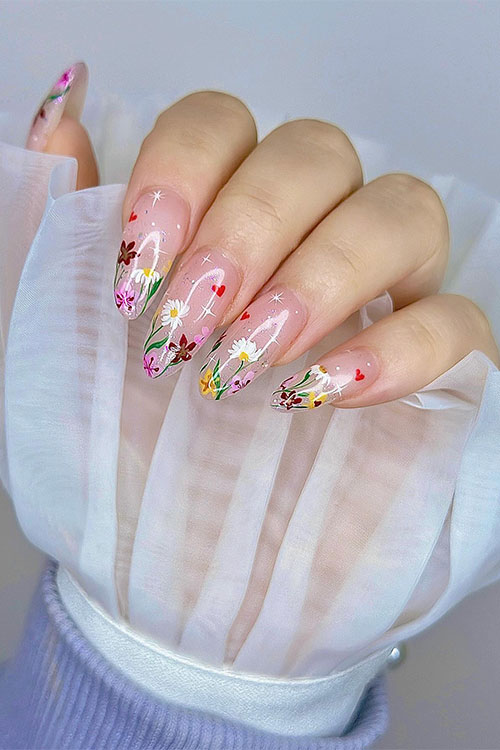 Long almond-shaped nude nails adorned with different flower colors on the nail tips besides, tiny white stars and red hearts