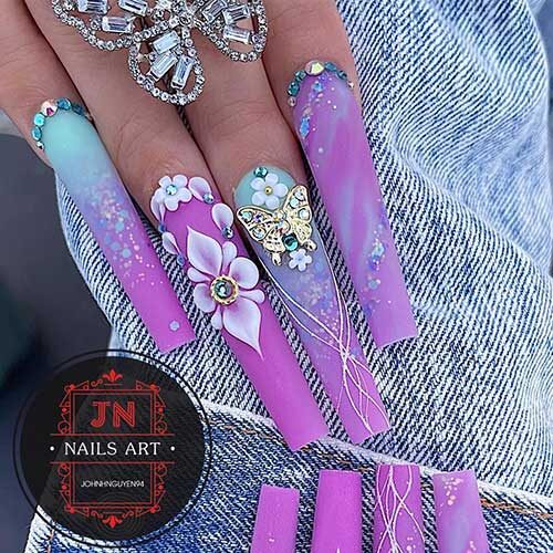 Long coffin-shaped matte purple and light blue nails with 3d flowers, rhinestones, and glitter