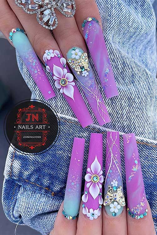 Long coffin-shaped matte purple and light blue nails with 3d flowers, rhinestones, and glitter