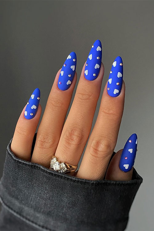 Matte cobalt blue Valentine's Day nails adorned with white heart shapes and rhinestones