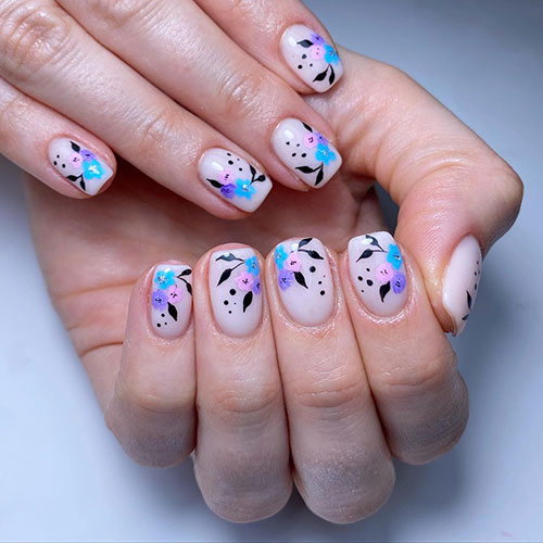 Short flower spring nails over nude base color and feature pastel flowers in blue, pink, and purple nail colors with black leaves