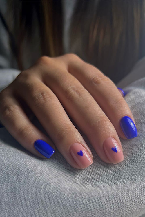 Short royal blue nails with two accent nude nails adorned with a royal blue heart shape on each accent nail