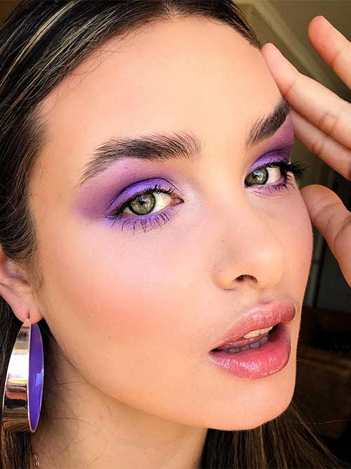 Striking purple eyeshadow highlights a woman's green eyes, complemented by rosy blush, glossy lips, and stylish purple hoop earrings. Striking purple eyeshadow highlights a woman's green eyes, complemented by rosy blush, glossy lips, and stylish purple hoop earrings.