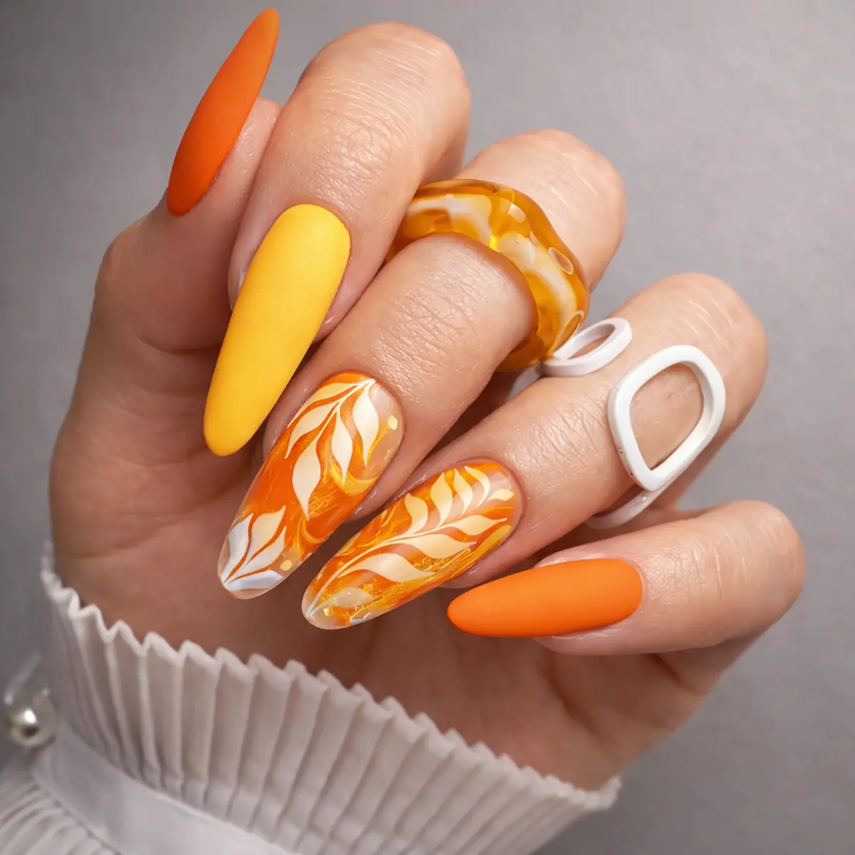 vibrant neon nails in matte tangerine orange and sunny yellow on almond-shaped tips, featuring elegant marble accent nails with white leaf art and gold swirls.