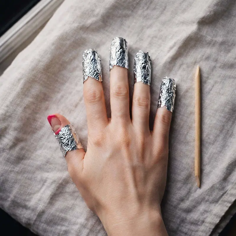 Flat-lay close-up of a woman's hand with all ten fingertips wrapped in small aluminum foil squares during a gel nail removal soak, with an orange stick placed beside the hand on a white linen surface