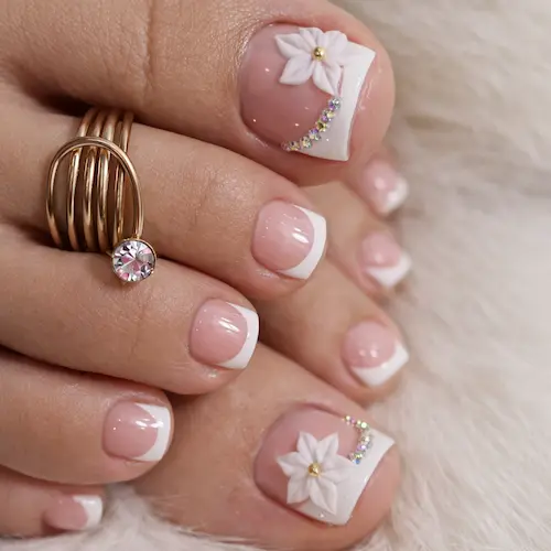 Detailed view of white flower press-on toenails featuring a pink nude base, French tips, and 3D floral designs with rhinestones.